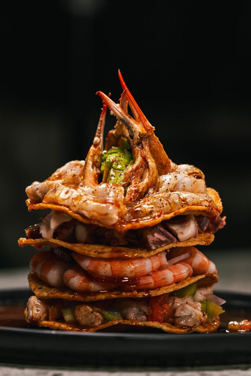 A stack of crisp tostadas layered with shrimp, octopus and avocado, crowned with a whole crab claw, lit on a black studio ground